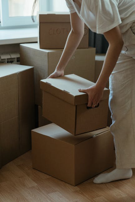 A person wearing light-colored clothing is inside a room, carefully lifting a medium-sized cardboard box, which is part of a larger collection of moving boxes stacked against a window. The boxes vary in size, some labeled with handwritten text such as 'CLOTHES,' and are open or closed with packing materials like bubble wrap or tissue paper visible. The individual appears to be in the process of packing or unpacking during a home relocation, with the boxes positioned on the wooden floor near the window area, which lets in natural light. Several boxes are arranged on the floor, with larger boxes supporting smaller ones, indicating an organized packing process. The image captures the typical scene of furniture transport and packing involved in house removals, with elements like cardboard boxes and careful handling suggesting the organized logistics of a professional moving service. The setting emphasizes the domestic environment and move-related activities, connecting to the services provided by Movers Stockwell in the realm of removals and move management.