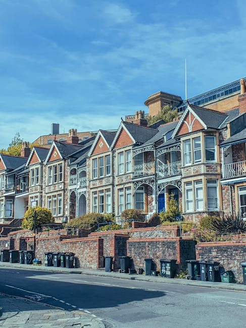 A row of Victorian-style terraced houses with decorative iron balconies and large windows, situated on a sloped street in Stockwell. The houses have brick facades with some featuring painted woodwork and ornate detailing around the windows and balconies. In front of the properties, a brick wall separates the houses from the pavement, which is lined with black wheelie bins. The sky above is clear with a few wispy clouds, and a large modern building can be seen in the background. This residential setting is typical of urban house streets in London, with the image capturing the exterior environment where home relocation and furniture transport may take place, reflecting the context of professional removals and packing services provided by Movers Stockwell.